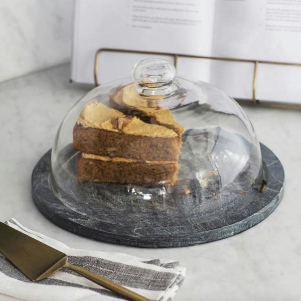A round, dark grey raw slate cake stand topped with a clear glass dome covering two generous slices of coffee and walnut cake on a marble worktop next to a striped cloth napkin and a gold cake slice.