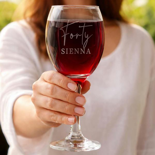 A hand holding a large, elegant wine glass filled with red wine. The glass is custom engraved with "Forty" in a delicate script font and "SIENNA" in bold uppercase letters. A soft-focus outdoor background highlights the crystal-clear glass.