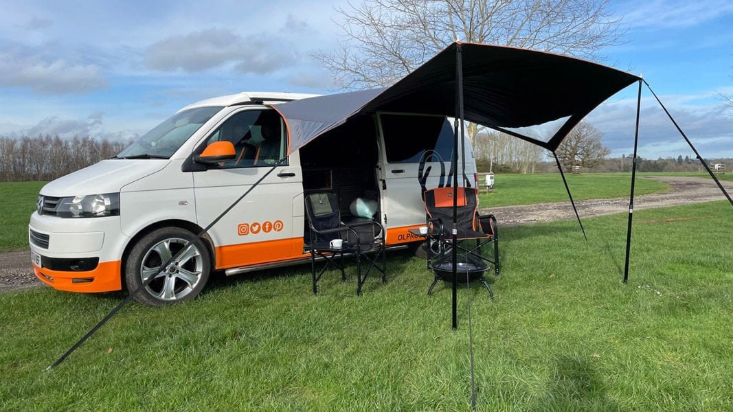 An image of the OLPRO Modern Shade Canopy attached to a Volkswagen T5 with some camping furniture displayed.