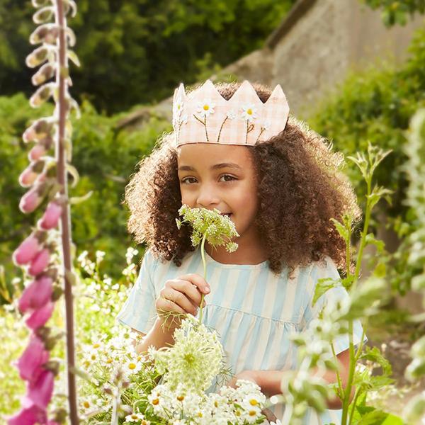 A young girl wearing a dusty pink gingham fabric crown with embroidered daisies while smelling a white flower in a summer garden.