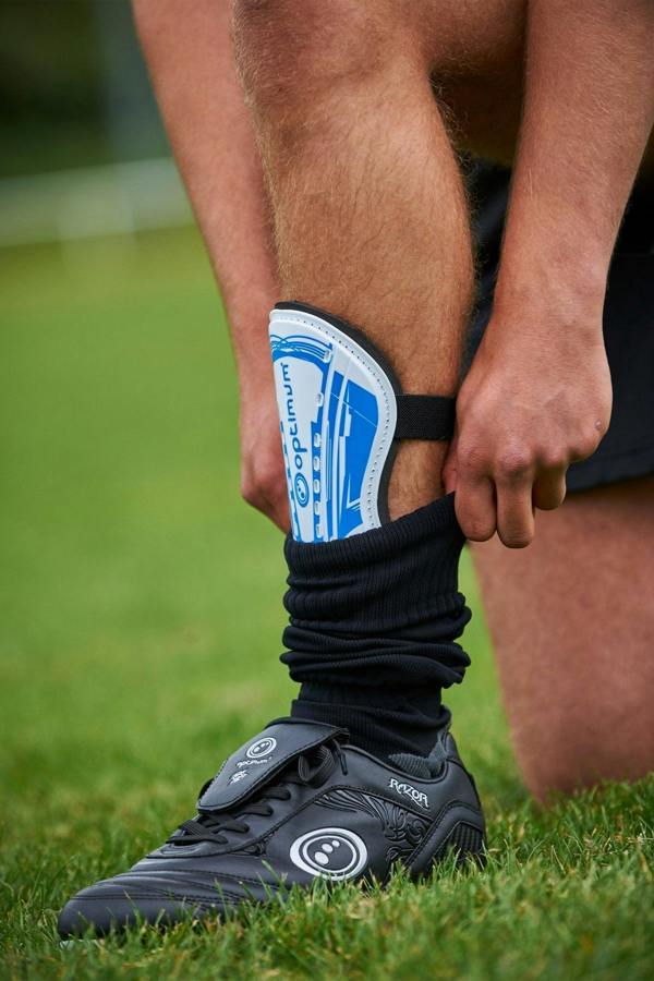 Player on grass wearing blue and white Optimum Velocidade shin guard over black sock with black football boots - Optimum