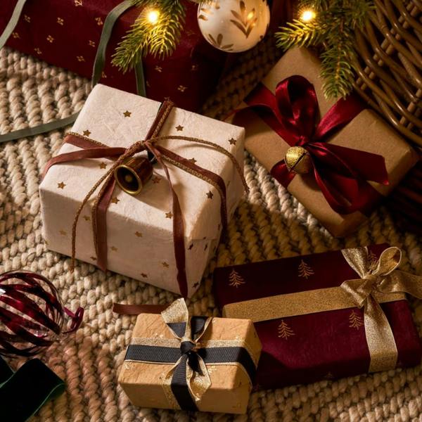 Selection of Christmas presents wrapped in berry red, warm white, and truffle cotton wrapping paper decorated with gold foil trees and stars.