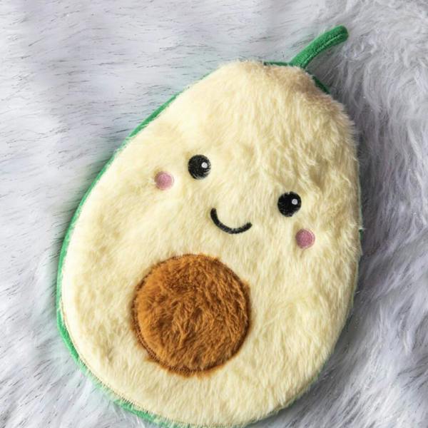 An overhead view of the Avocuddle Avocado Hot Water Bottle featuring a smiling embroidered face and a raised plush brown seed center, resting on a white faux-fur shag rug.