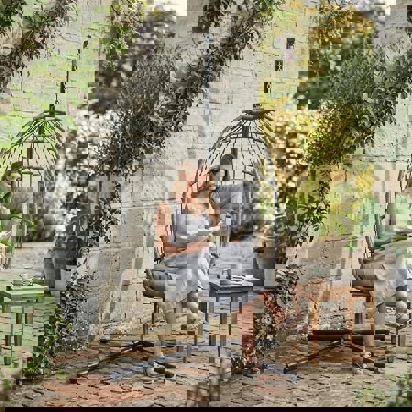 Grey rattan hanging egg chair with plush cushions, featuring a sleek design, set in a lush garden with a woman reading a book