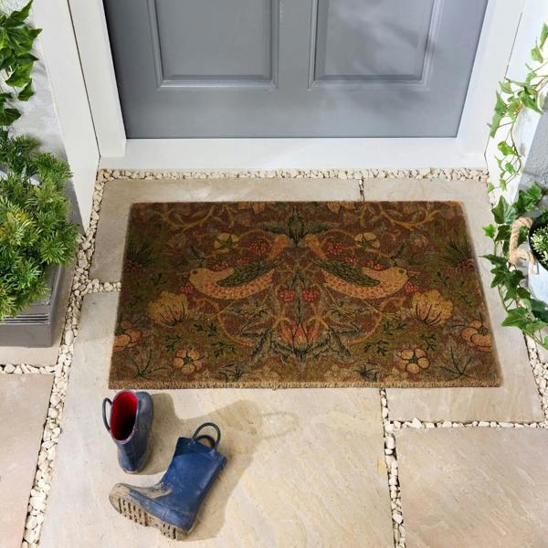Decorative doormat with floral pattern in front of a gray door, with boots and plants on either side.