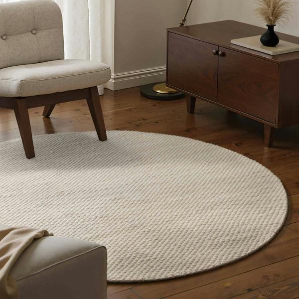 Round beige rug in a living room with wooden furniture and a beige armchair.