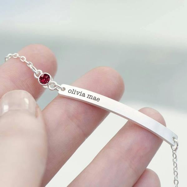 A hand holding a silver Personalised Birthstone Swarovski Crystal Bracelet custom engraved with the name olivia mae next to a small round red crystal.