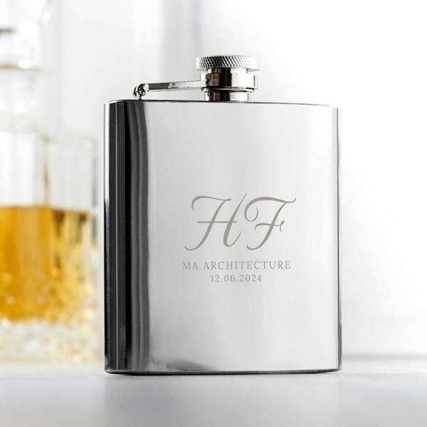 A sleek polished stainless steel graduation hip flask engraved with initials HF, MA Architecture, and a ceremony date, standing on a light table.