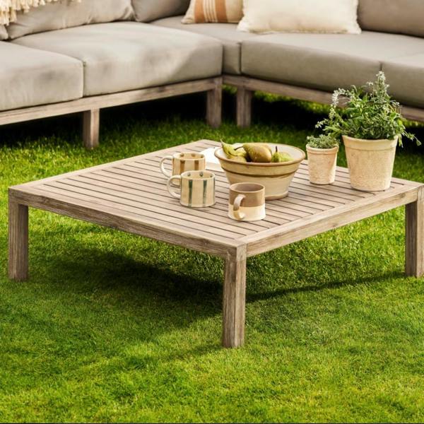 Square whitewash acacia wood coffee table styled with drinks and fruit on a garden lawn.
