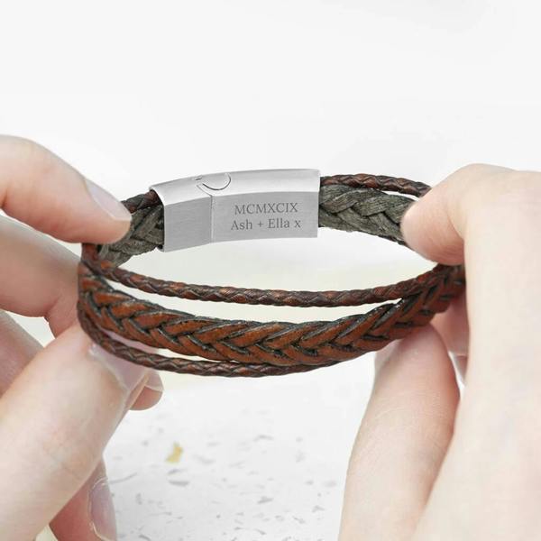 Hands holding open a personalised layered brown leather men's bracelet showing a silver clasp engraved with "MCMXCIX" and "Ash + Ella x".