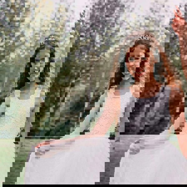 Young girl in a white dress with a tiara, standing outdoors with trees in the background