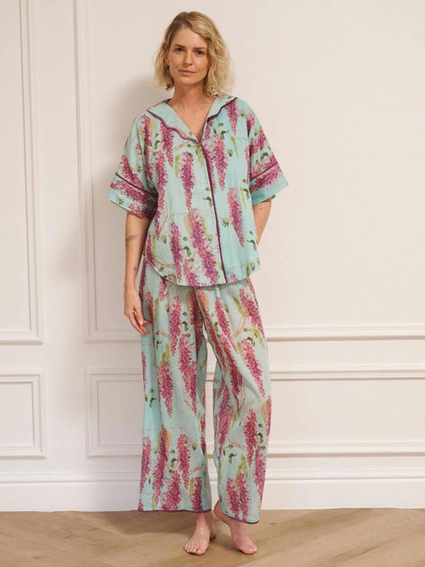 Woman wearing duck egg blue cotton gauze oversized pyjamas with pink wisteria floral print, short-sleeve button-through top and wide-leg trousers, standing barefoot indoors.