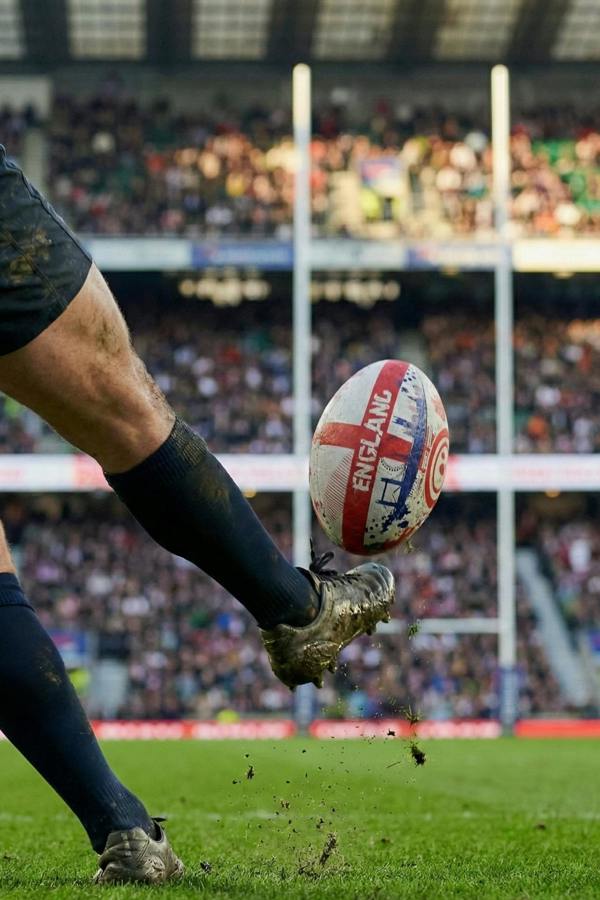 Player kicking a white and red Optimum England rugby ball in a packed stadium, ball mid-air - Optimum