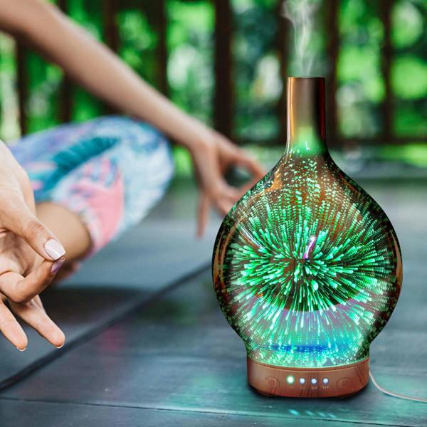 A person meditates cross-legged beside the glowing Rio ELLA Aroma Diffuser, Humidifier, and Night-Light, which emits soothing vapor and colorful light for a relaxing outdoor ambiance.