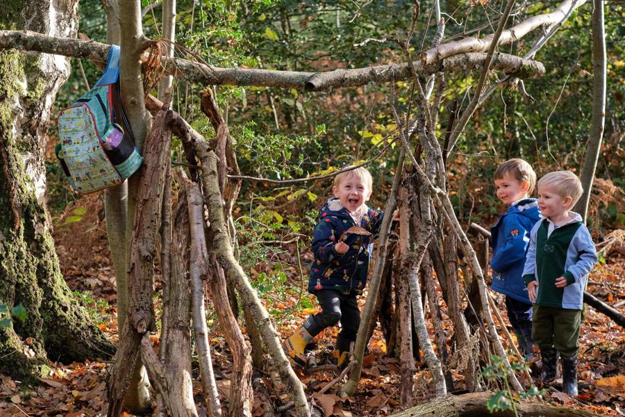 toddlers playing around a wooden den in a forest setting with a more toddler meal in the foreground.