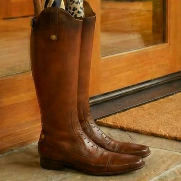 A quirky brown hand-painted polyresin umbrella stand shaped like a pair of leather riding boots, sitting on a stone floor by a wooden glass panel door holding umbrellas.