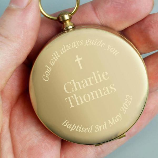 A close-up of a gold-tone, brushed metal compass held in a person's hand. The compass is engraved with a cross and the fixed text "God will always guide you," along with personalised name and date details.