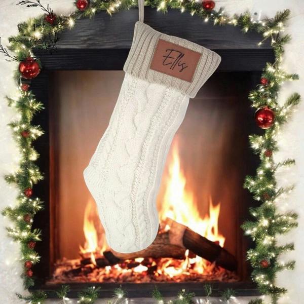 Personalised cream cable-knit Christmas stocking with an engraved faux leather patch reading Ellis hanging in front of a glowing fireplace hearth.
