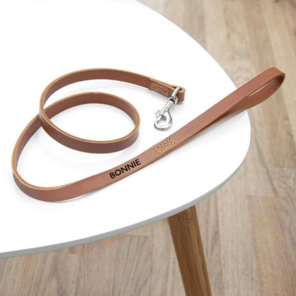 A personalised classic brown leather dog lead embossed with the name BONNIE in black foil, coiled on a white wooden table.