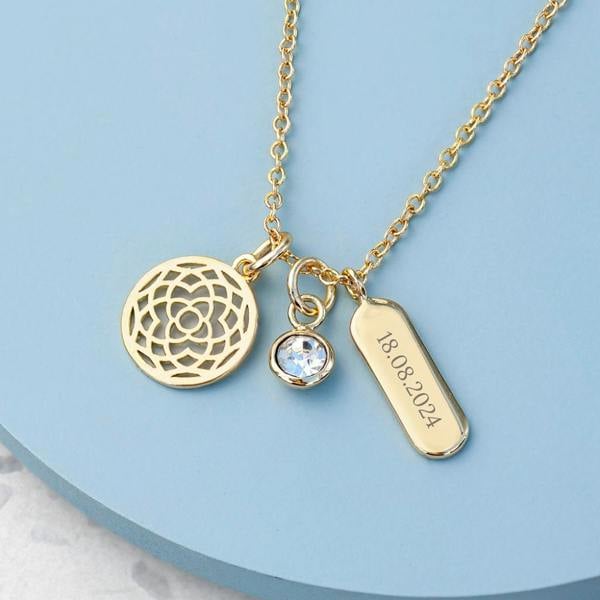 A close-up of a gold personalised mandala charms necklace featuring an openwork circular mandala charm, a bezel-set clear crystal charm, and an engraved gold bar displaying the date '18.08.2024' on a light blue round background.