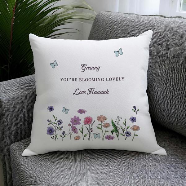 A personalised white cushion printed with a wildflower border and the text 'Granny You're Blooming Lovely Love Hannah' resting on a grey fabric armchair.