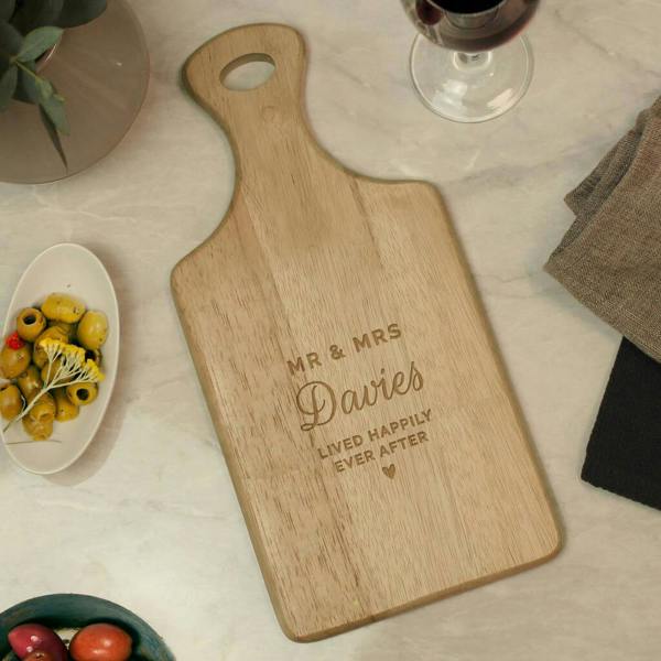 A top-down view of a rectangular wooden paddle board with a handle on a marble surface, custom-engraved with the words Mr and Mrs Davies Lived Happily Ever After, placed next to a bowl of olives and a glass of red wine.