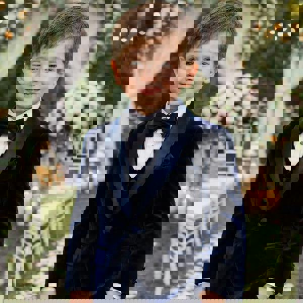 Young boy in a navy velvet suit standing in front of floral decorations at an outdoor event.