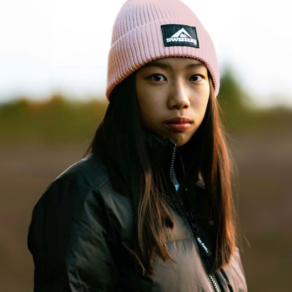 Young woman wearing a simple beanie hat knitted in a chunky single rib in a flamingo pink shade. There is a SWIMZI badge sewn to the front of the deep turn back headband.