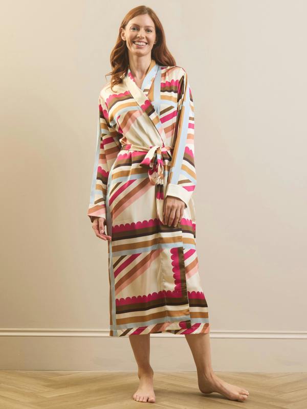 A woman modelling a long-sleeve wrap dressing gown robe with a colourful geometric stripe pattern in pink, brown, beige, and light blue, standing against a plain beige wall.