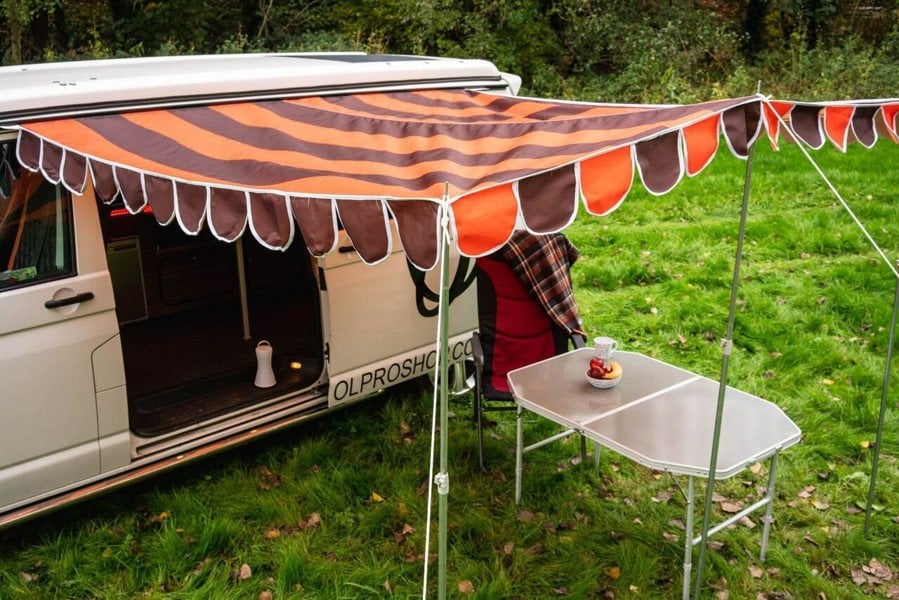 A picture of the OLPRO Orange and Brown Campervan Canopy that is attached to a Volkswagen Camper Van, with some camping furniture underneath it.