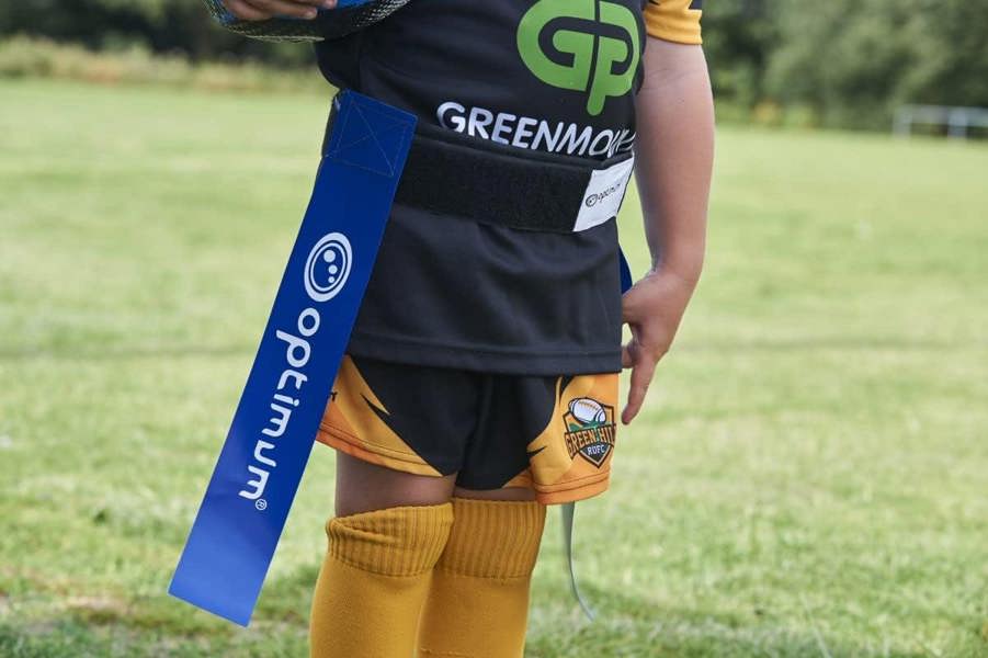 Blue Optimum tag rugby flag worn on a young player's waist over black and orange kit, on grass