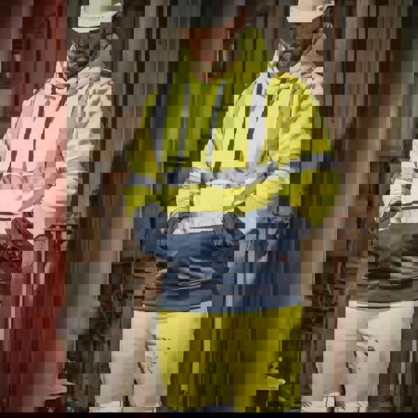 Scruffs High Visibility Mens Hi-Viz Work Pull Over Hoodie, yellow and navy high-visibility hoodie with reflective strips, being worn by a construction worker in a safety helmet inside a workshop.