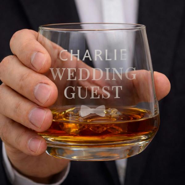 A heavy-bottomed whisky tumbler held by a man in a suit. The glass is engraved with "Charlie Wedding Guest" and a small bow tie motif, filled with amber-coloured spirits.