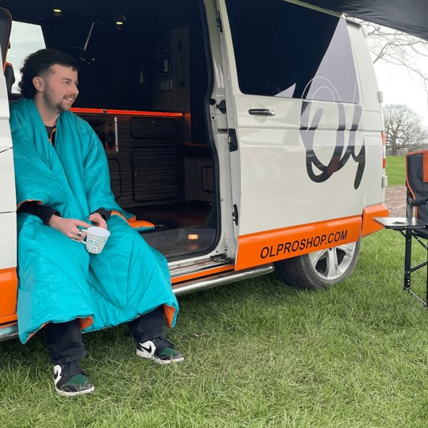 A man sits on the step of a VolksWagen T5 campervan in his insulated reversible blanko blanket, whilst drinking from a mug.