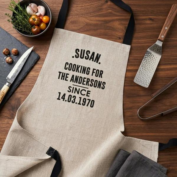 An overhead flat lay photograph of a personalised linen cooking apron, custom printed with '.Susan. Cooking for the Andersons Since 14.03.1970', styled on a dark wood background alongside an artisanal knife, black slate, bowl of tomatoes and garlic, a hammered spatula, and metal tongs.