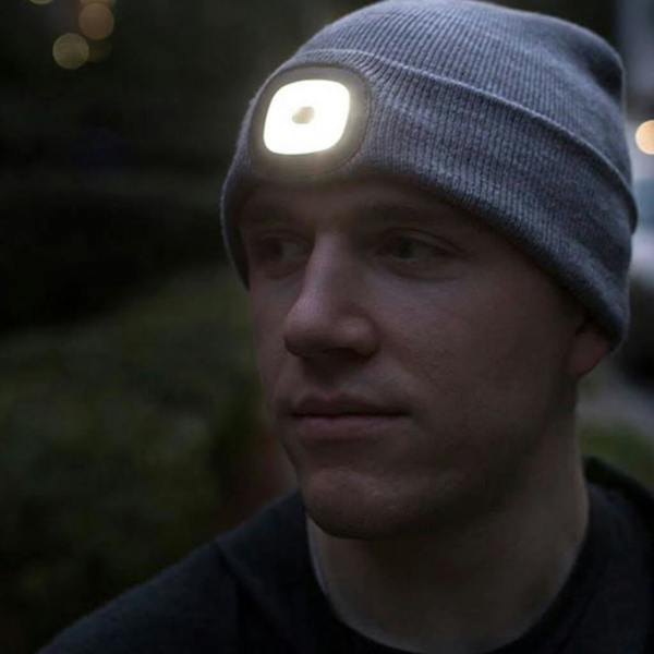 A person wearing a grey, knitted beanie hat in a dark outdoor setting. A bright LED light is integrated into the front of the hat, illuminating the area in front of the wearer.