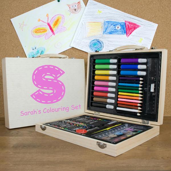 A 63-piece personalised children's colouring set in a wooden case printed with a large pink letter S and Sarah's Colouring Set, open on a desk displaying art supplies alongside drawing artwork.