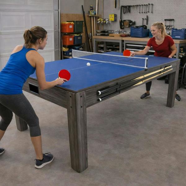 7-in-1 games table with a blue surface, sturdy grey wood frame, and two players enjoying a game of table tennis in a garage setting