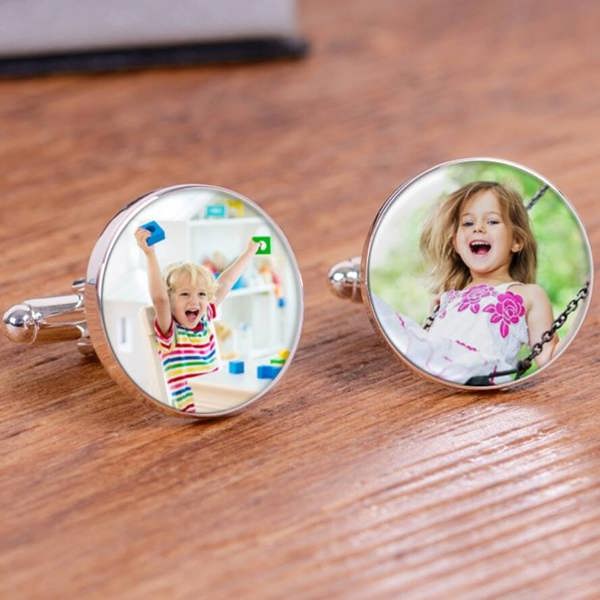 A close-up shot of a pair of round silver finish photo cufflinks resting on a warm wooden surface, displaying vibrant images of a young boy playing with blocks and a young girl laughing on a swing.