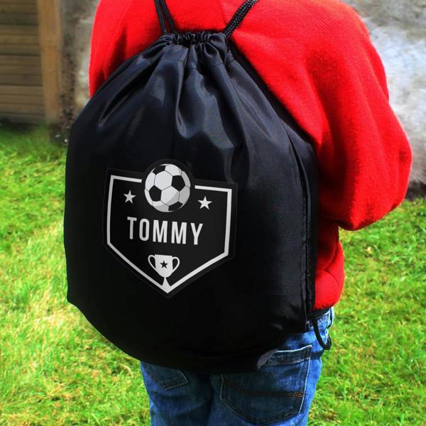 A child wearing a red jumper and jeans from behind, holding a black personalised drawstring kit bag with a football crest and the name 'Tommy'.