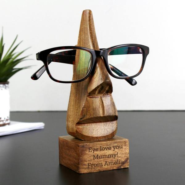 Personalised nose-shaped acacia wood glasses stand on a black desk with reading glasses, custom engraved for Mummy.