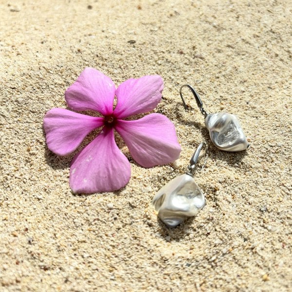 Drop earrings featuring electroplated silver, irregular shells, photographed on a sand next to an exotic flower.