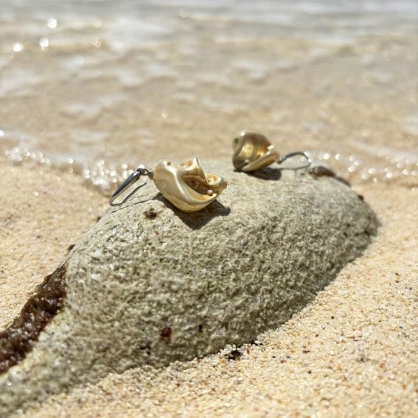 Golden shell earrings by LENAE displayed on a beach.
