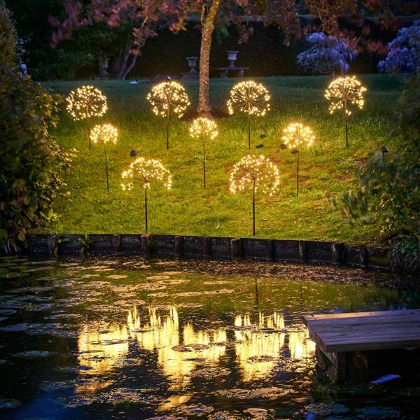 Multiple warm white solar dandelion stake lights illuminating a grassy slope at dusk, reflecting beautifully in a garden pond in the foreground.