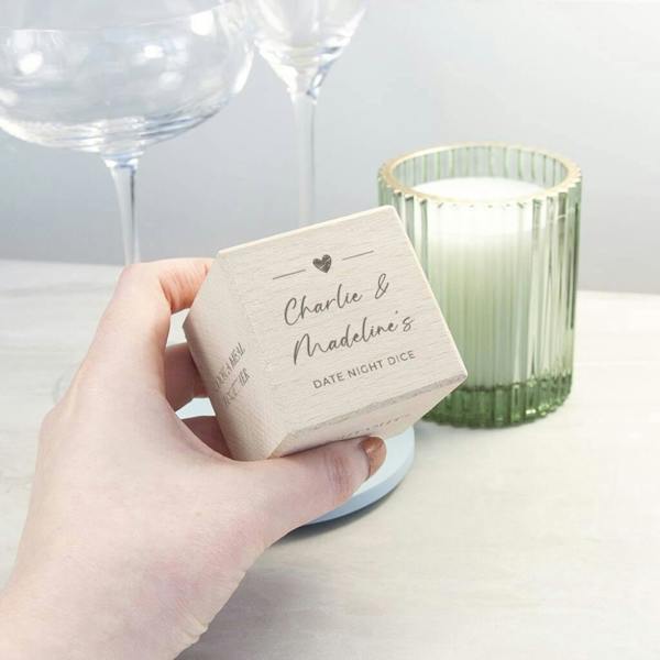 A close-up of a hand holding a square wooden date night die on a marble table, personalised with the text "Charlie & Madeline’s Date Night Dice" and a heart icon, with a lit candle and wine glasses blurred in the background.
