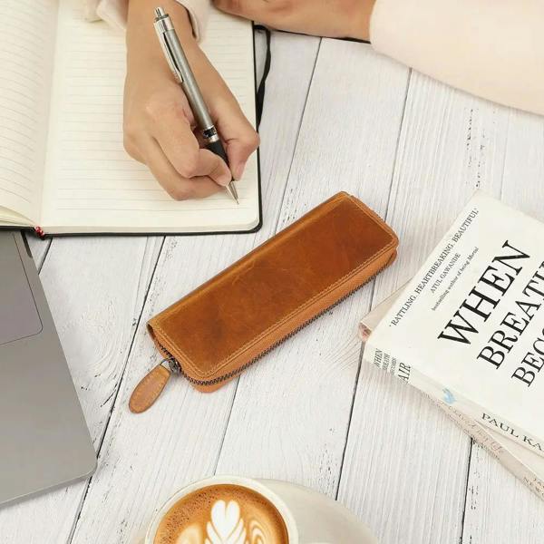 Tan leather pen case on a wooden surface with a notebook, pen, and coffee.