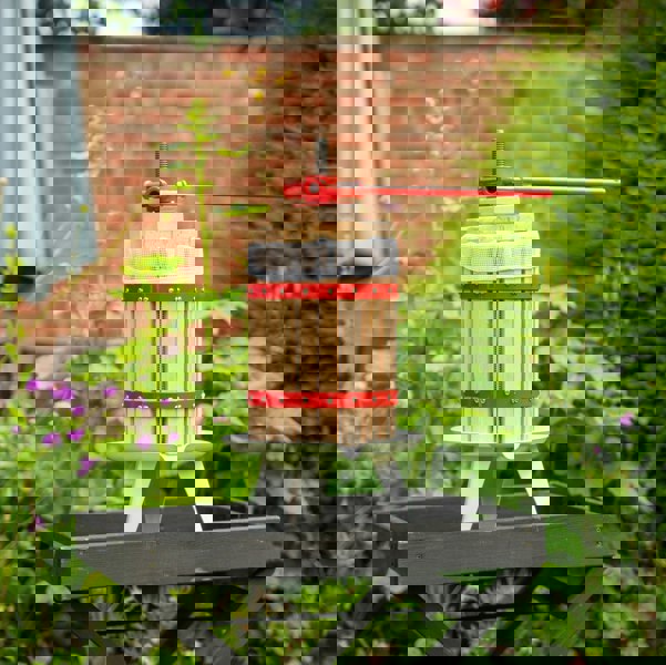 KuKoo 18 Litre Fruit Press with wooden slats, red metal accents, and a sturdy base, set in a vibrant garden with flowers