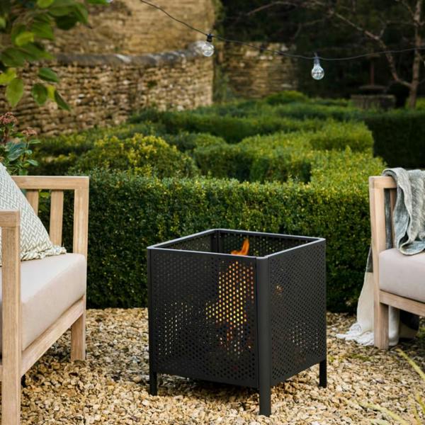 Industrial matte black square mesh fire pit with a lit log fire, styled on a gravel patio between two wooden garden armchairs.