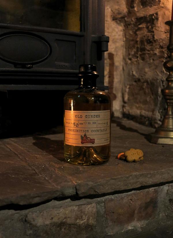 A bottle of Old Ginger Prohibition Cocktails filled with amber liquid, set on a rustic stone hearth next to a small gingerbread cookie in a cozy, dimly lit fireplace setting.