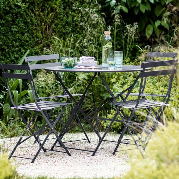 A French-inspired carbon dark grey metal bistro set featuring a round dining table and four folding chairs on a gravel patio surrounded by lush green foliage.